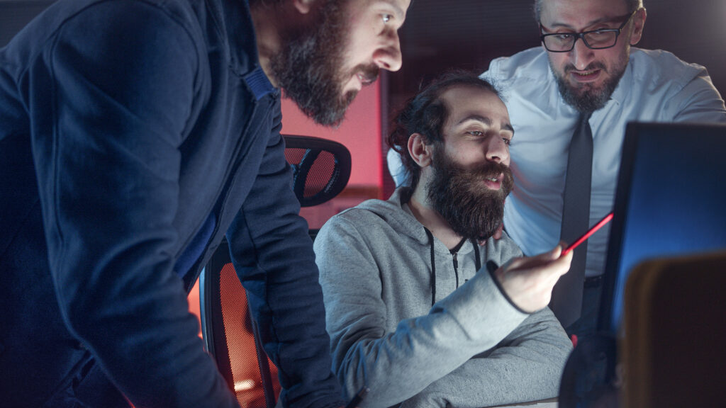 Three male colleague workers collaborating in office talking while looking at computer screen together at workplace